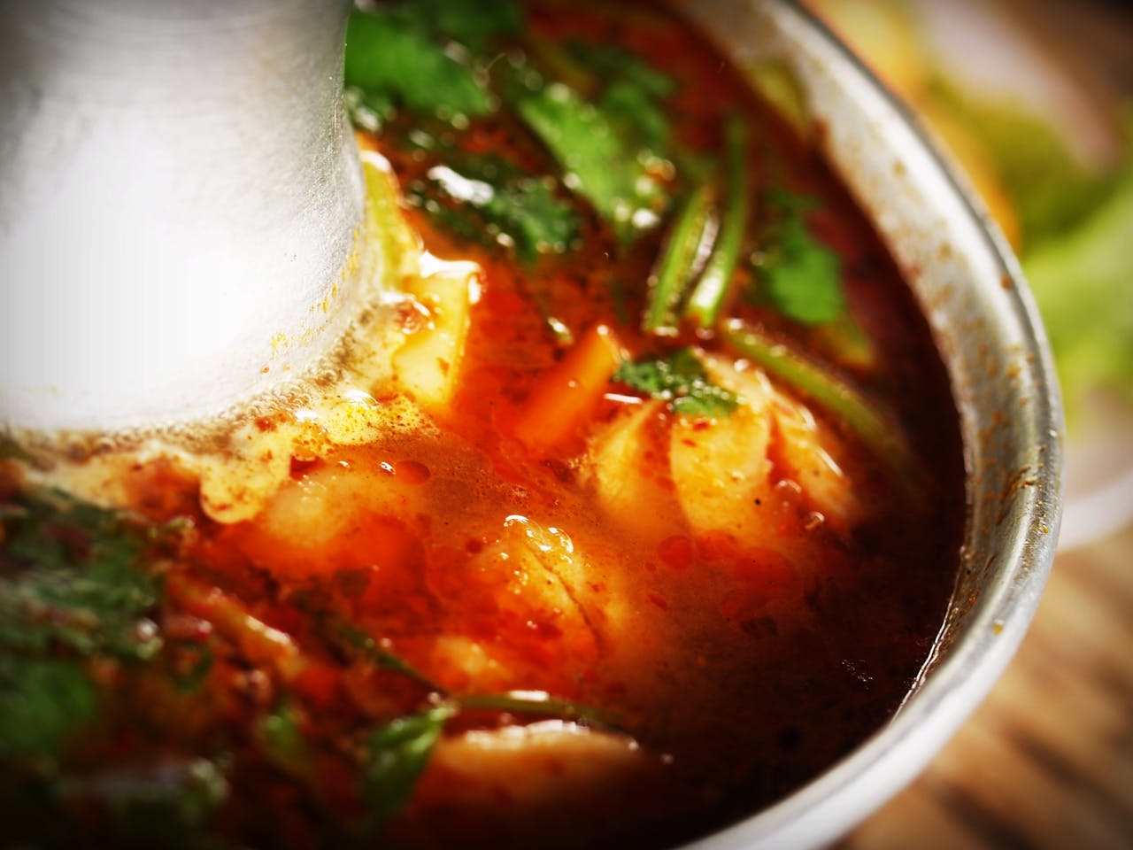 gallery-4 Close-up of flavorful Thai Tom Yum soup with aromatic herbs in a metal bowl.