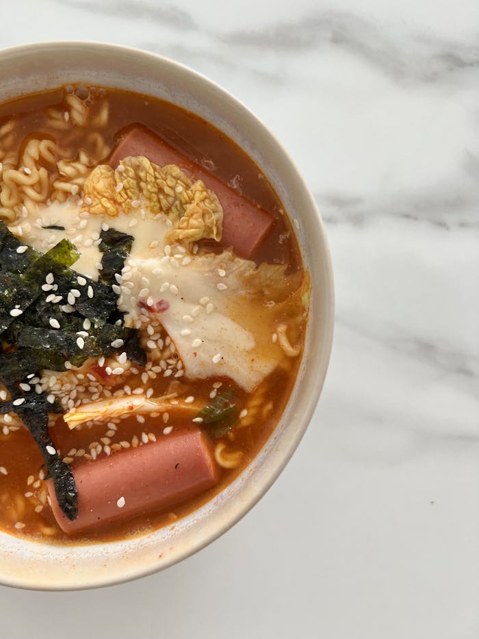 gallery-5 A close-up of a flavorful Korean ramyeon with cabbage, seaweed, and sausage.